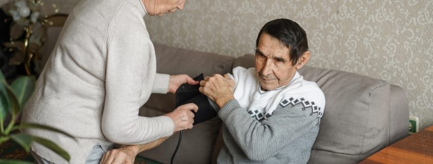 Vanem naine mõõdab diivanil istuva vanema mehe vererõhku. Mees hoiab oma kätt väljaspoole, samal ajal kui naine reguleerib vererõhumansetti tema ülemise käe peal. Nad on elutoas siseruumides.