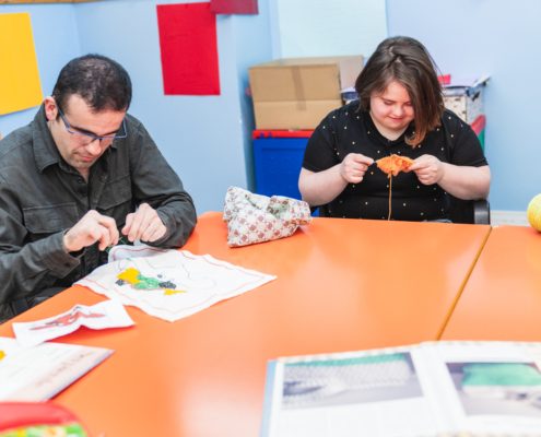 Mees ja naine istuvad oranži laua ääres ja tegelevad käsitööprojektidega, neid ümbritsevad käsitöötarbed ja avatud raamat - võib-olla arutavad nad üheskoos töötades teenused või sotsiaalkindlustus.