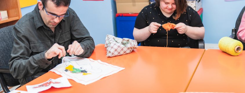 Mees ja naine istuvad oranži laua ääres ja tegelevad käsitööprojektidega, neid ümbritsevad käsitöötarbed ja avatud raamat - võib-olla arutavad nad üheskoos töötades teenused või sotsiaalkindlustus.