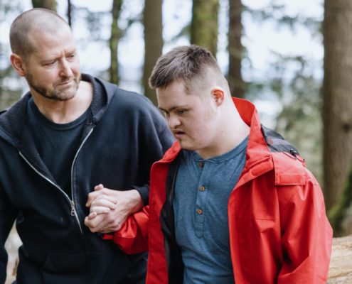 Kaks meest kõnnivad koos metsas. Üks punases jakis mees vaatab alla, teine, kellel võib olla kogemusi hooldusvajadusega ringide eest hoolitsemisel, hoiab õrnalt tema käest kinni ja vaatab teda hoolitseva pilguga.