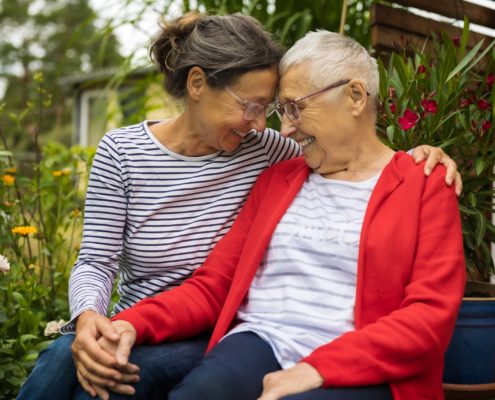Kaks naist istuvad tihedalt koos verandal, naeratavad ja puudutavad otsaesist. Nad hoiavad käest kinni, jagades omastehoolduse hetke; ühel on punane jakk ja prillid, teisel on pruunid juuksed punnis ja seljas triibuline särk.
