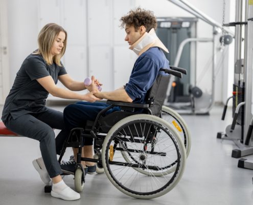 Naine abistab hooldusvajadusega inimene ratastoolis, kes kannab kaelarihma, käteharjutuse tegemisel teraapiapalliga taastusravi jõusaalis.