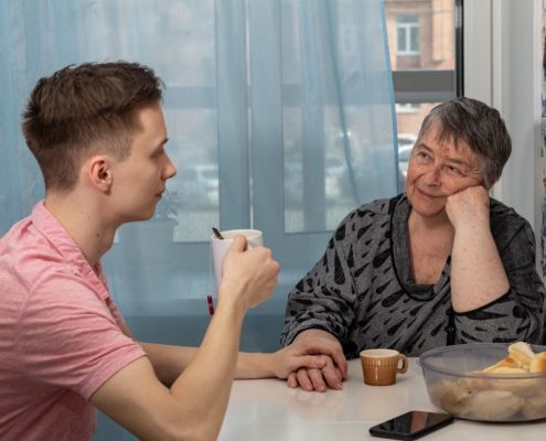 Noormees ja vanem naine istuvad laua taga käest kinni hoides. Keset toitu, tassi ja telefoni räägivad nad vaikselt kaashoolduskoormusest - nende tähelepanelikes žestides ja soojas ühenduses peegeldub hoolduse ühine raskus.