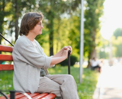 Lühikeste juustega ja prillidega vanem naine istub pargis punasel pingil, kepi käes, vaatab kõrvale ja naudib päikesepaistelist päeva. Õues viibimine võib vaimne tervis, nagu näha puud ja inimesed eemal, väga kasuks tulla.