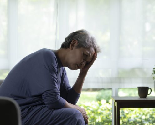 Lühikeste hallide juustega vanem inimene istub siseruumides diivanil, kummardudes ettepoole, üks käsi otsaesisel, näib olevat stressis või mures - hetk, mis peegeldab muret vaimne tervis. Ees on laud, millel on tass ja potitaim, valgusvihkab läbi suurte akende.