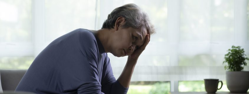 Lühikeste hallide juustega vanem inimene istub siseruumides diivanil, kummardudes ettepoole, üks käsi otsaesisel, näib olevat stressis või mures - hetk, mis peegeldab muret vaimne tervis. Ees on laud, millel on tass ja potitaim, valgusvihkab läbi suurte akende.