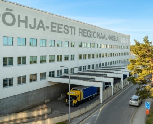 Suur valge hoone, mille fassaadile on kirjutatud PÕHJA-EESTI REGIONAALHAIGLA, millel on mitu akent, kollane ja sinine veoauto ning puudega vooderdatud sõidutee-paik kukkumise ennetamine foorum 22. septembril.