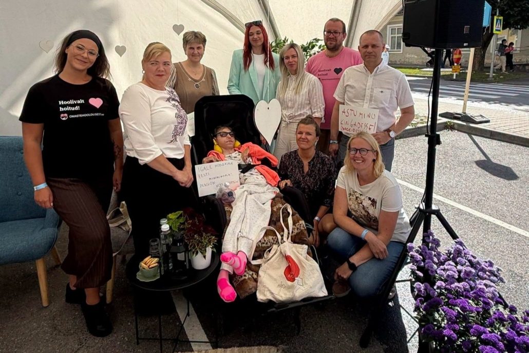 Rühmapilt üritustelgis, kus üheksa inimest seisavad ja istuvad käsitsi kirjutatud siltide taga. Kesksel kohal ratastoolis istuva inimese ees on silt tekstiga "Igale hooldajale Eesti pension, miinimumpalk hooldajatele". Paremal seisab mees, kelle sildil on kiri "Astu minu kingadesse!". Roosas särgis mehe särgil on tekst "Hoolivalt hoitud!" ja veebiaadress omastehooldusest.ee.