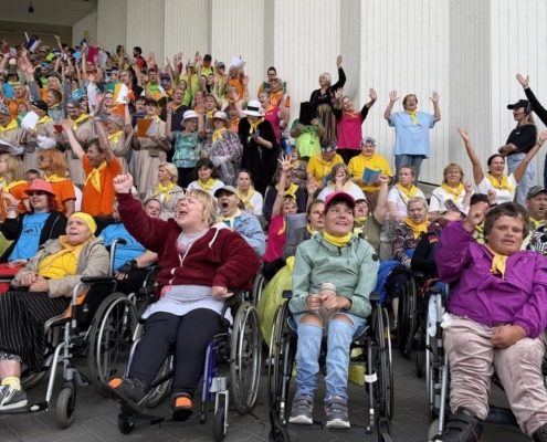 Ratastoolikasutajate rühm rõõmustab rusikaid tõstes suure rahvahulga ees, kes kannavad kollaseid salle ja tähistavad üritust õues.