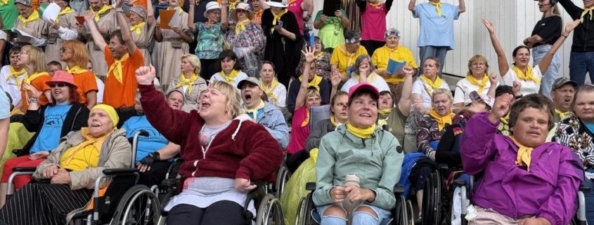 Ratastoolikasutajate rühm rõõmustab rusikaid tõstes suure rahvahulga ees, kes kannavad kollaseid salle ja tähistavad üritust õues.