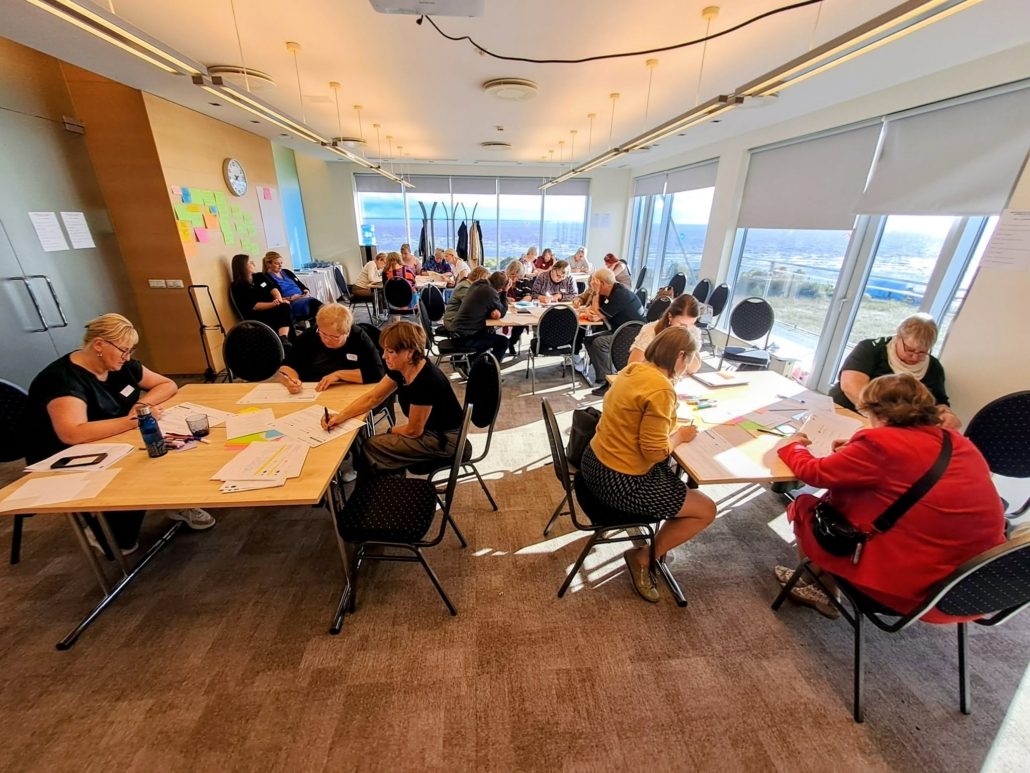 Seminaril osalejad täidavad laudade taga töölehti ja kirjutavad värvilistele kleepmärkmetele; suurtest akendest avaneb vaade merele.