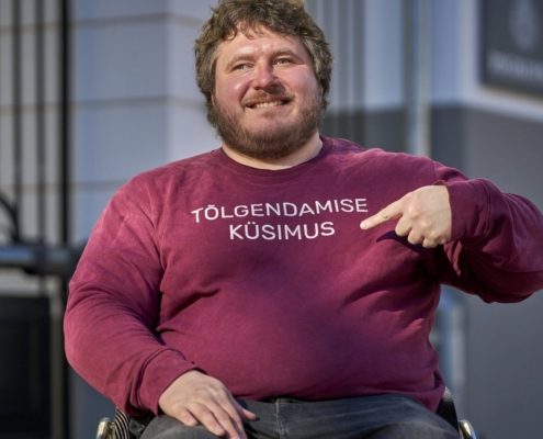 Naeratav habemega mees ratastoolis osutab sõrmega oma bordopunasel dressipluusil olevale tekstile: „TÕLGENDAMISE KÜSIMUS".