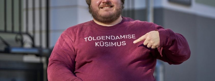 Naeratav habemega mees ratastoolis osutab sõrmega oma bordopunasel dressipluusil olevale tekstile: „TÕLGENDAMISE KÜSIMUS".